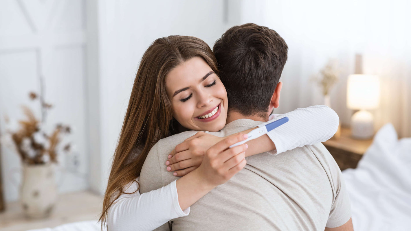 Eine glückliche Frau umarmt ihren Mann und hält einen positiven Schwangerschaftstest in der Hand